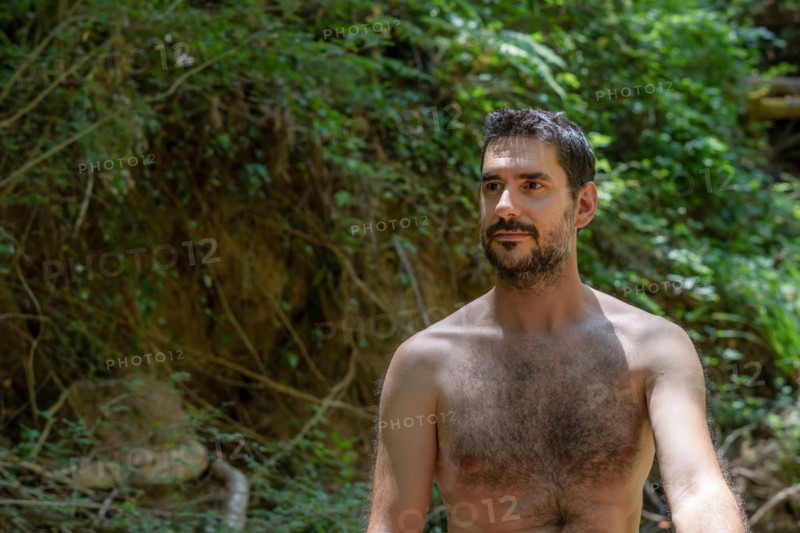 Shirtless young man sitting on the bank of a river with the forest in the background