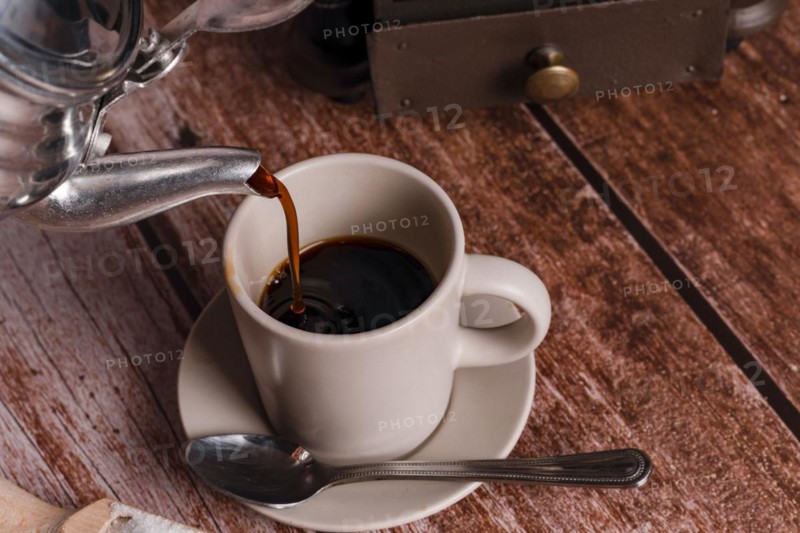 Metal vase pouring coffee into a ceramic cup on a wooden table
