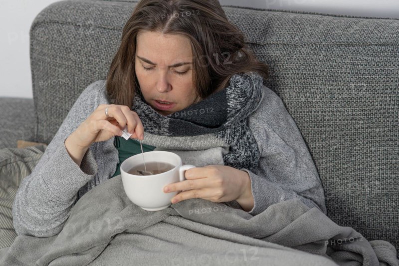 Sick woman covered with a blanket drinking an infusion on the couch in her home