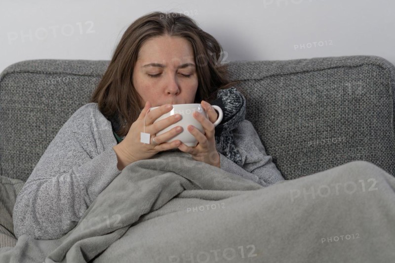 Sick woman covered with a blanket drinking an infusion on the couch in her home