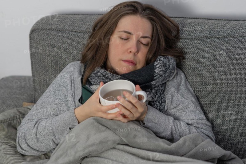 Sick woman asleep on couch covered with a blanket with a cup of tea in her hand