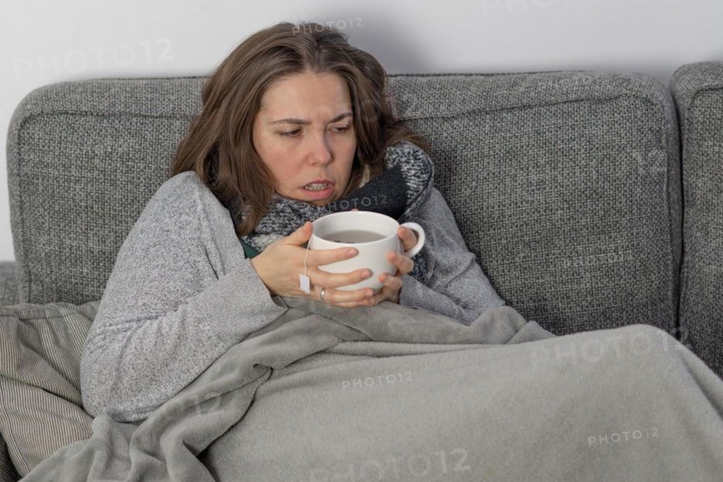 Sick woman covered with a blanket drinking an infusion on the couch in her home