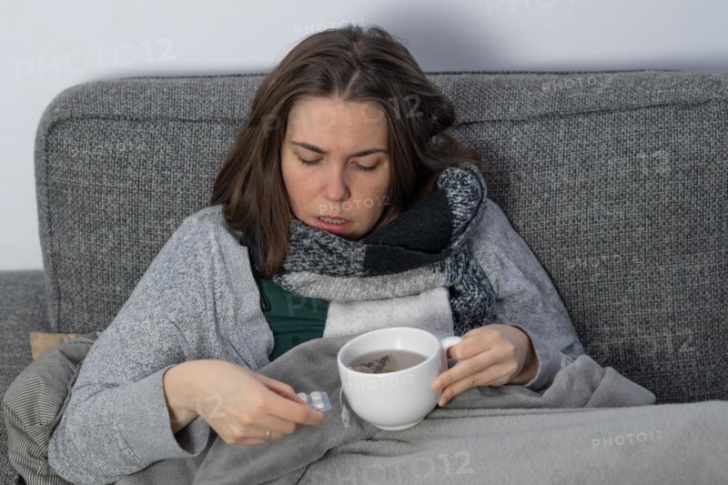 Sick woman covered with a blanket drinking an infusion on the couch in her home