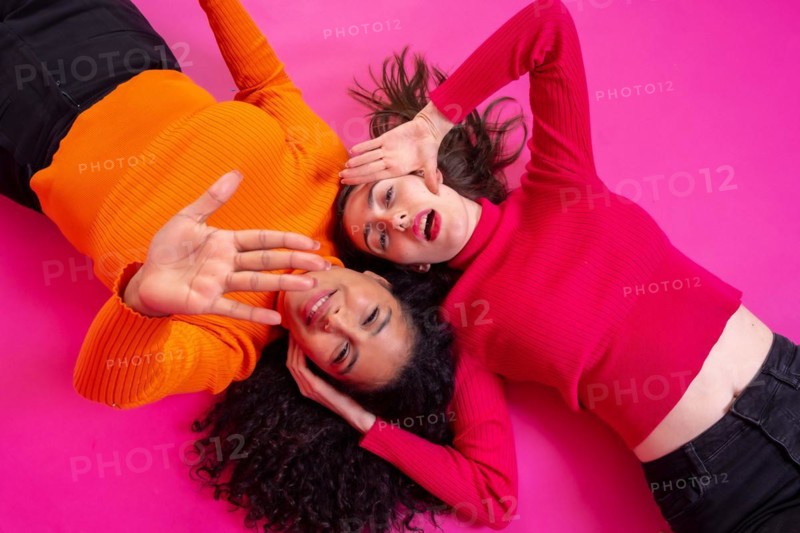 Two multi-ethnic women lying down making stop gesture with hands over pink background