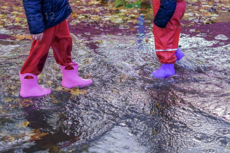 Two children walking through a puddle of water with their boots on a rainy day