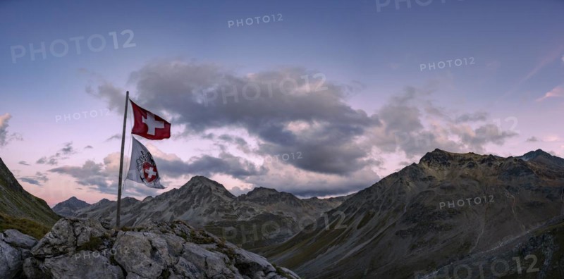 Swiss flag and flag of the SAC with cloudy sky at blue hour and Engadine mountains