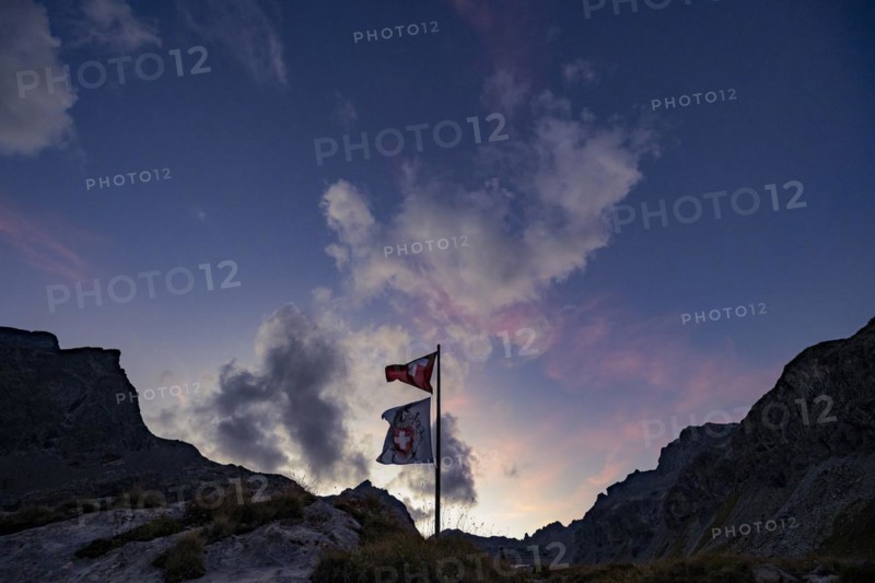 Swiss flag and flag of the SAC with cloudy sky at blue hour and Engadine mountains