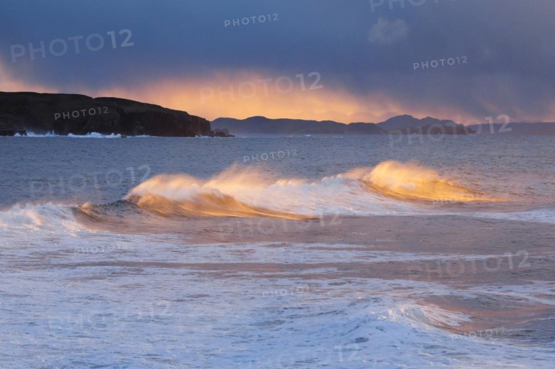 Large waves crash in a winter storm and the swirling spray is illuminated by the warm light of the evening sun