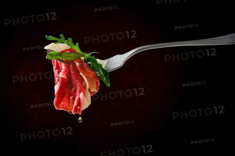 Piece of marbled beef carpaccio on fork with dripping olive oil over black background