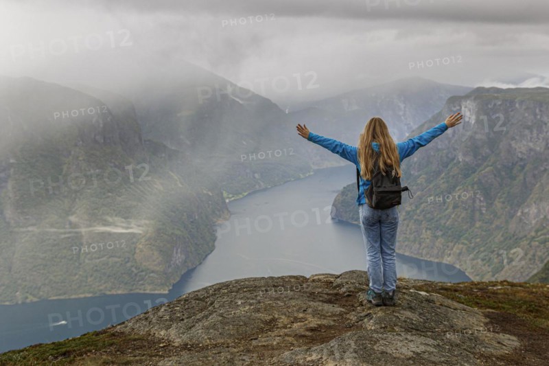 A girl stands on a mountain above the Aurlandsfjord in Norway and stretches her hands up in the air