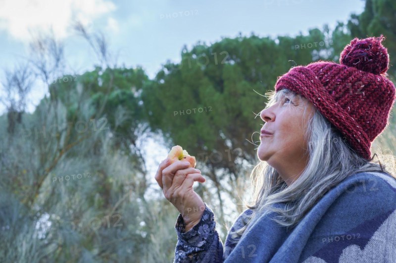 White-haired woman with red cap covered with a blanket eating an apple in the mountains