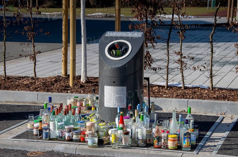 Bottles and jars stand in front of an overcrowded waste glass collection container
