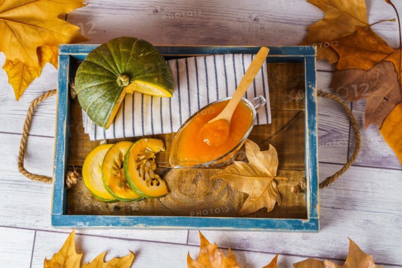 Top view of homemade pumpkin marmalade in a glass jar on a wooden tray with dried leaves