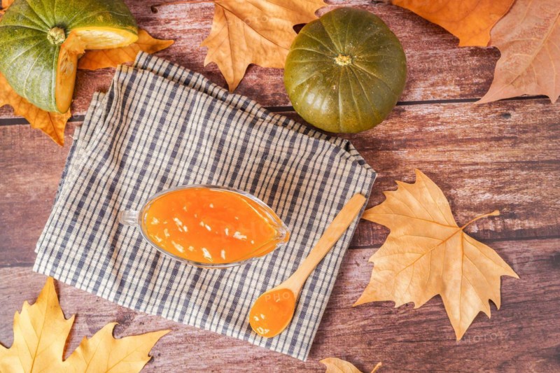 Homemade pumpkin marmalade in a glass jar with dried leaves on a white cloth on a wooden table