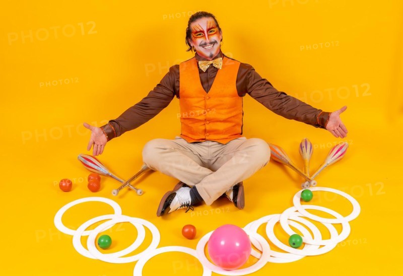 Portrait of a juggler in a vest and makeup sitting with the juggling objects on a yellow background