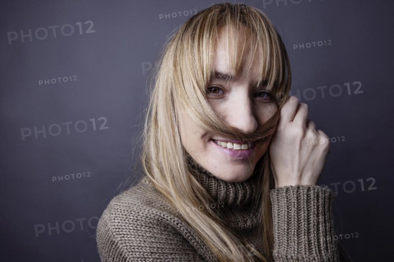Young woman with long blond hair makes a moustache out of a strand of hair and smiles at camera