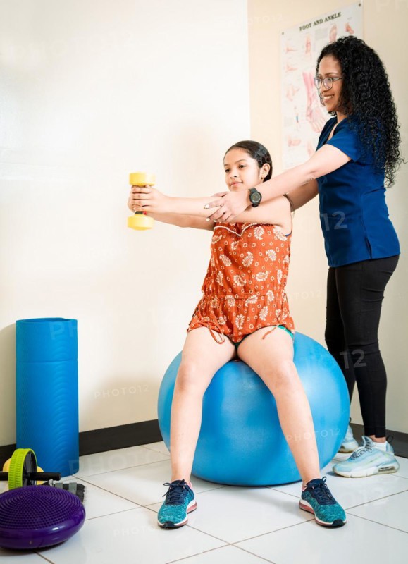 Physiotherapist with female patient holding hand dumbbell sitting on rehab ball. Physiotherapist helping female patient with hand weight sitting on fitness ball