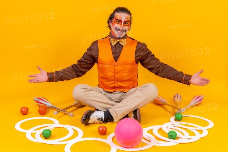 Juggler in a vest and with his face painted sitting with the juggling objects on a yellow background