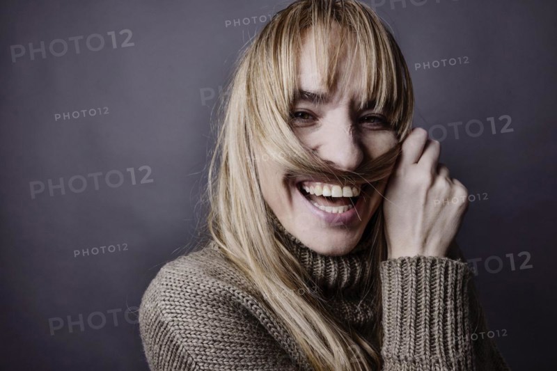 Young woman with long blond hair makes a moustache out of a strand of hair and smiles at camera