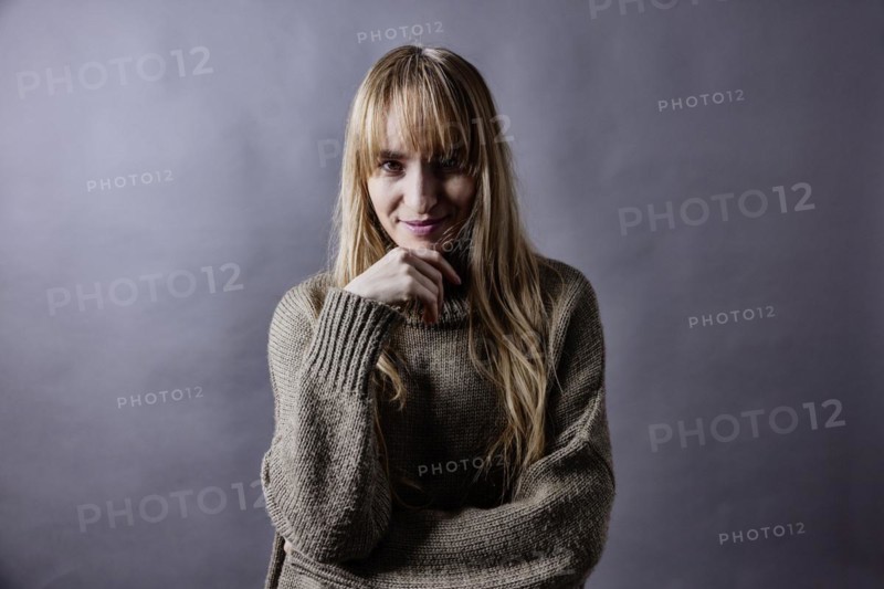 Woman with long blond hair and woollen jumper holds a hand to her chin and smiles at camera