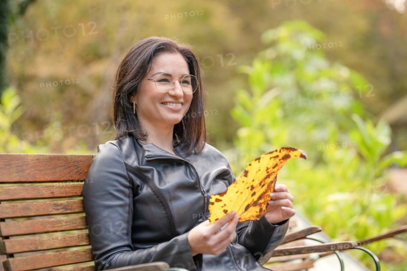 Pretty woman with glasses and leaf in hand sits on bench in park