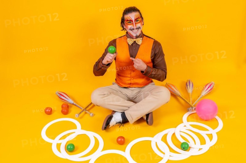 Portrait of a juggler in a vest and makeup sitting with the objects on a yellow background
