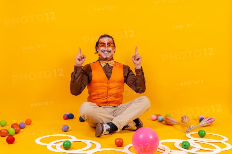 Juggler in waistcoat and with painted face sitting with the juggling objects on a yellow background