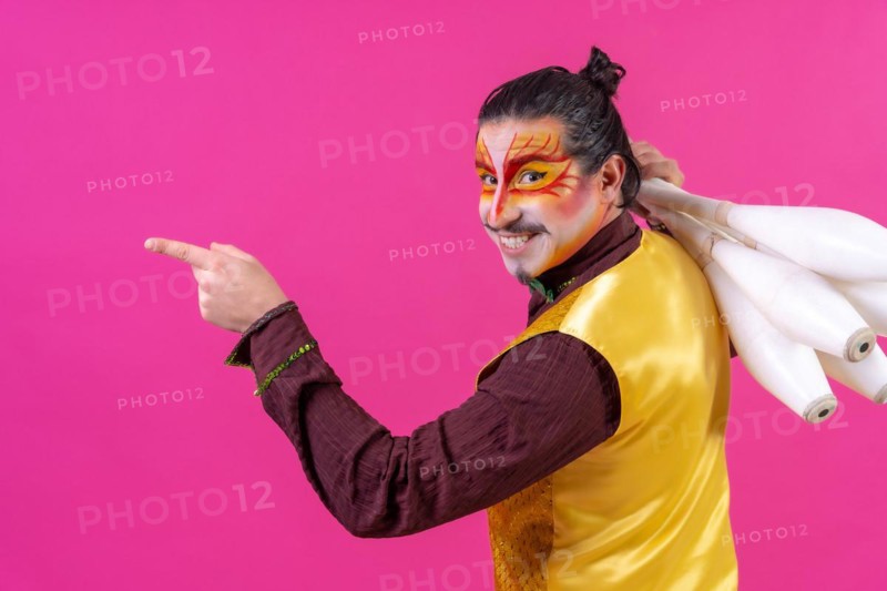 Juggler in a vest and with painted face juggling maces on a pink background