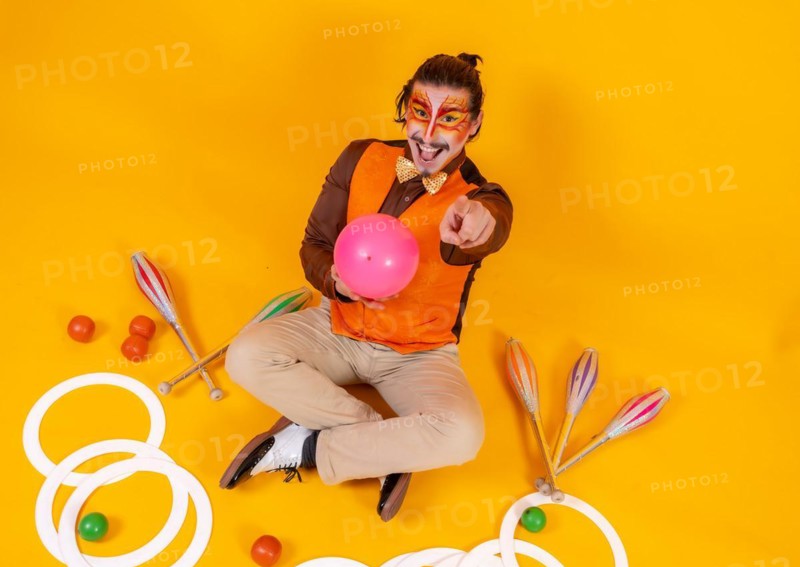Portrait of a juggler in a vest and makeup sitting with the juggling objects on a yellow background