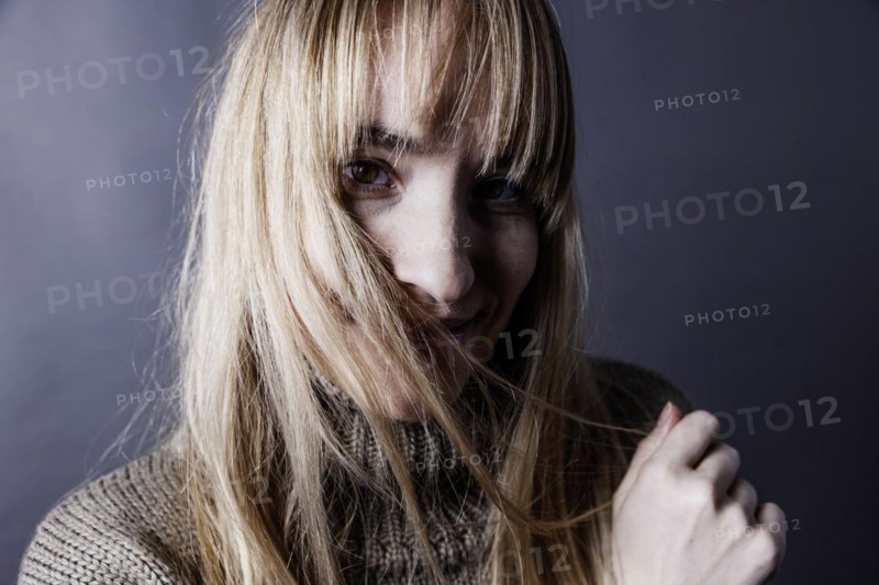 Young woman with long blond hair covers one half of her face with a strand of hair and looks saucily into camera