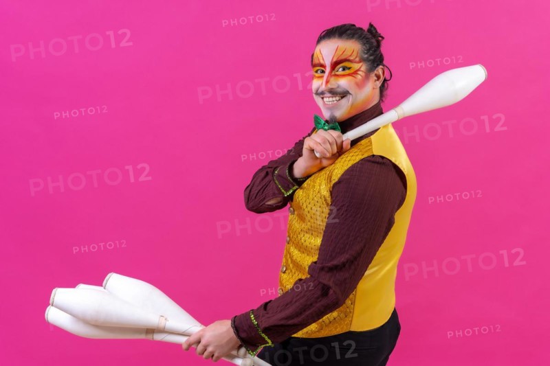Portrait of a juggler in a waistcoat and with painted face juggling maces on a pink background