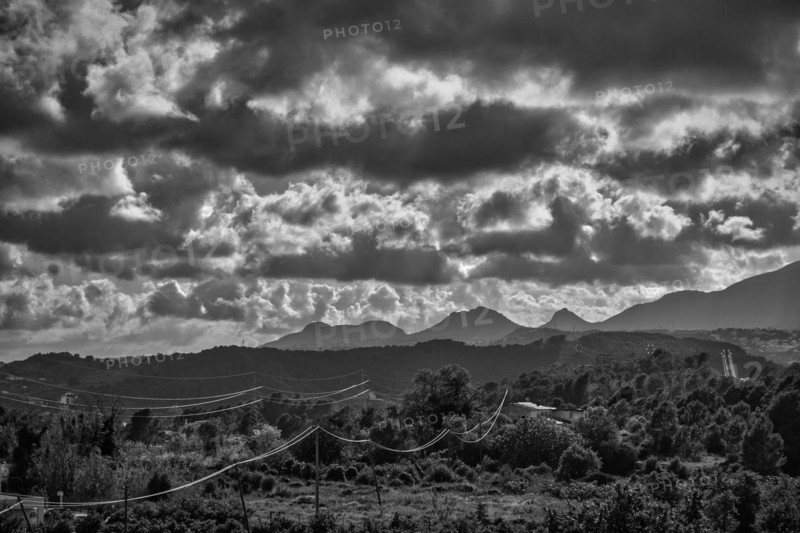 Dramatic sky over rugged mountain silhouettes and woods crossed by power lines in black and white in Spain