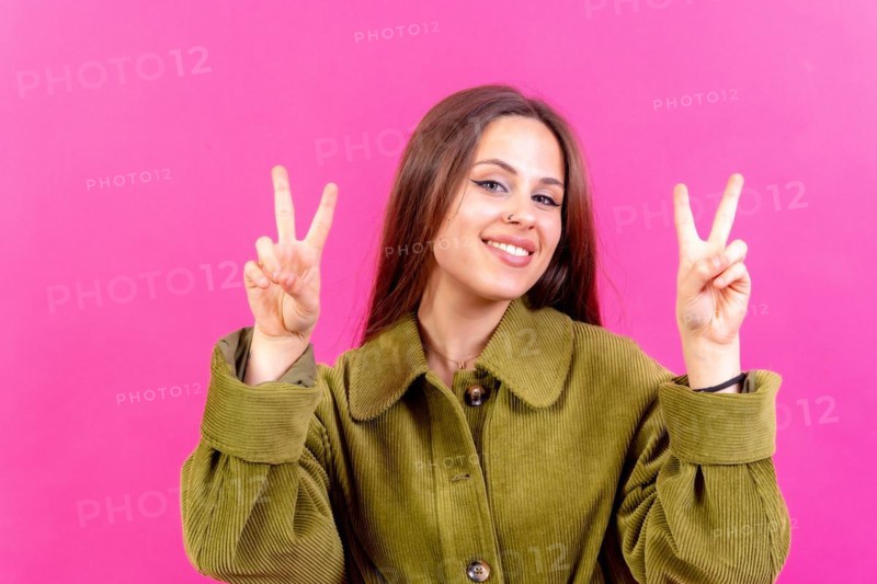 Young woman over pink background smiling and showing victory sign wearing green windbreaker