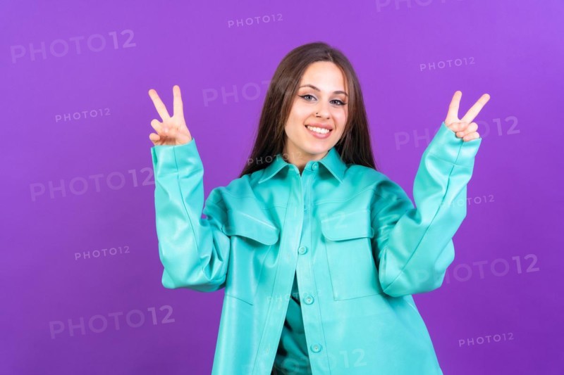 Young woman isolated on purple background smiling and showing victory sign wearing blue windbreaker