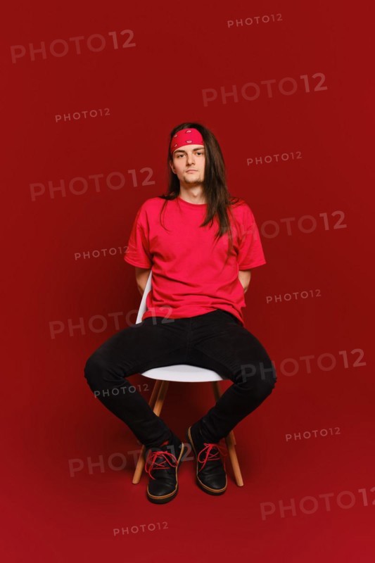 Young adult man in black and red casual clothes sits on chair with his arms behind back