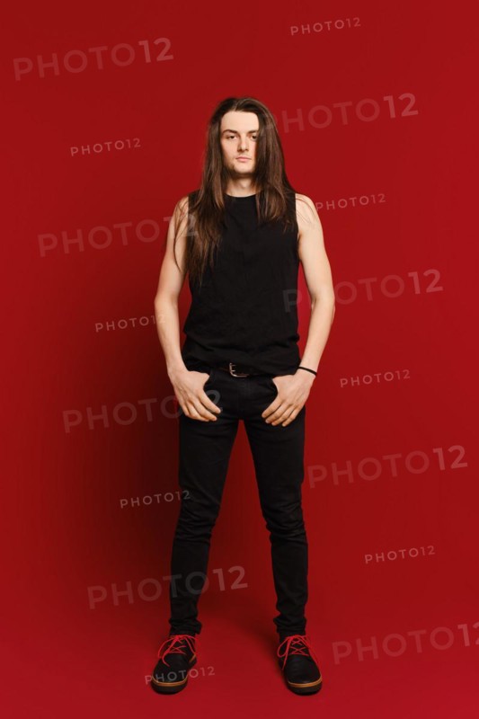 Full body indoor portrait of positive young man with long hair standing against red background