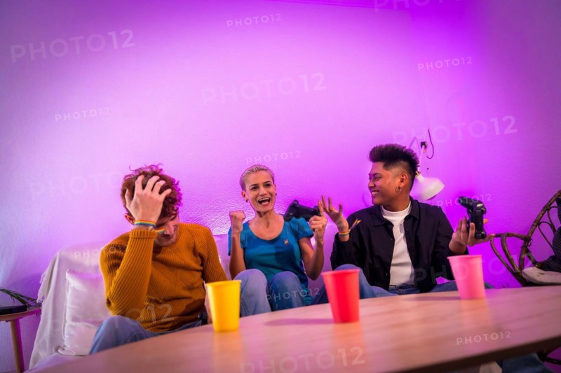 Group of young friends playing video games together on the sofa at home, purple led, woman winning