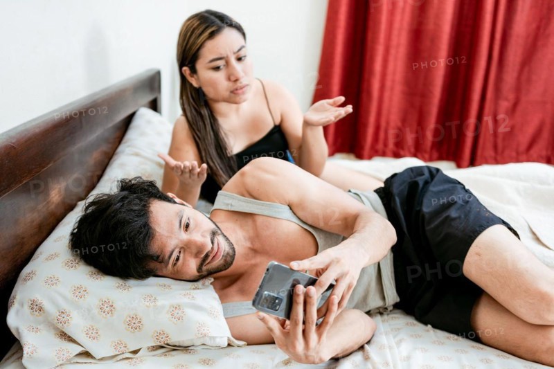 Cheating Husband Texting On Phone Ignoring Wife Lying In Bed. Husband with cell phone ignoring wife in bed. Annoyed woman with husband while holding cell phone in bed