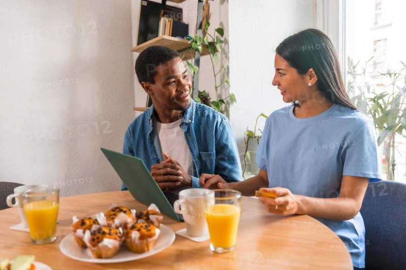 Multi-ethnic couple making purchase online with the computer while having breakfast, next to the window
