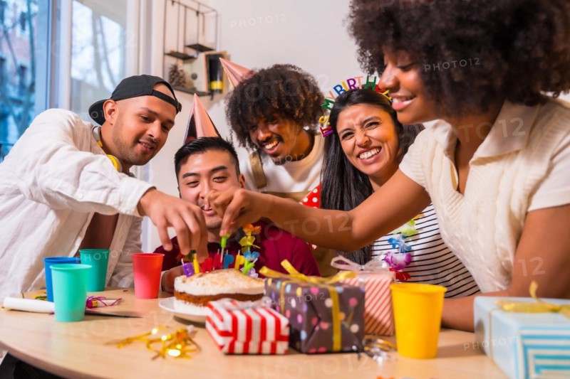 Multi-ethnic group of friends at a birthday party on the sofa at home with a cake and gifts