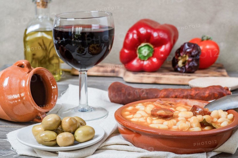 White beans stewed with bacon and chorizo in an earthenware casserole on a wooden table with all the ingredients of the recipe