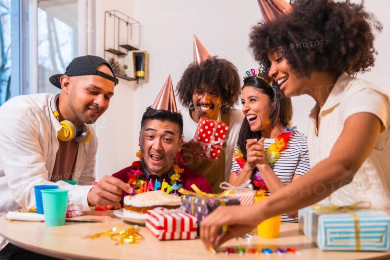 Multi-ethnic group of friends at a birthday party on the sofa at home with a cake and gifts
