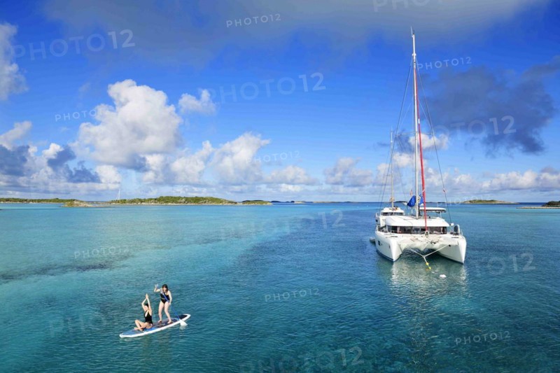 Women paddle SUP and do yoga in front of a sailing catamaran in the harbour of Warderick Wells