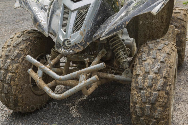 Black front rubber tires covered with dried mud on All Terrain Vehicle parked on gravel road surface