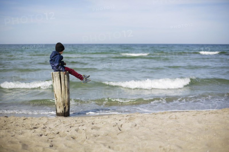 Symbolic photo on the theme of loneliness in childhood. A child sits alone on a wooden post by the sea. Arenshoop