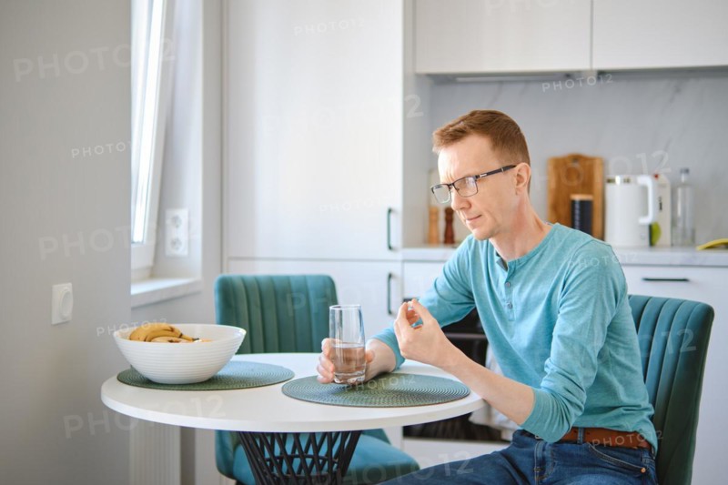 Middle aged man is sitting behind the kitchen table and holding a pill and a glass of water in hands