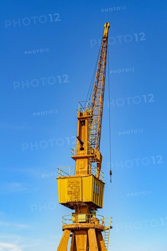 Old and obsolete yellow crane on the harbor pier lit by the afternoon sun with blue sky in the background.