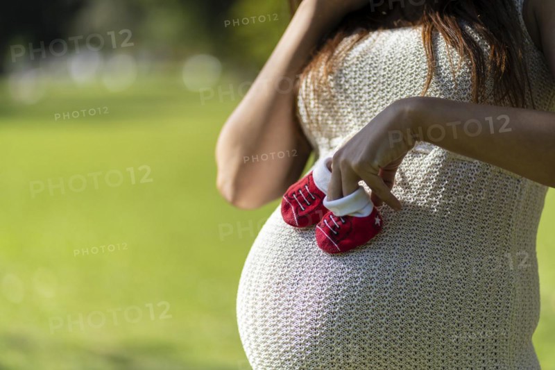 Pregnant latin woman holding baby shoes on her tummy in a park