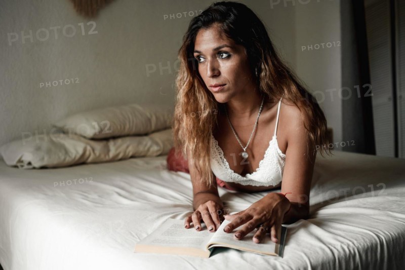 Young latina girl with long hair reading a book lying on her bed is distracted looking towards the window