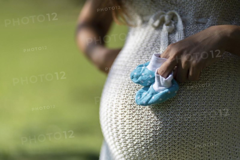 Pregnant latin woman holding baby shoes on her tummy in a park